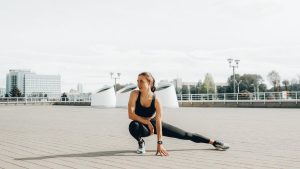 Young woman in black sportswear stretching outdoors in a sunny urban environment.
